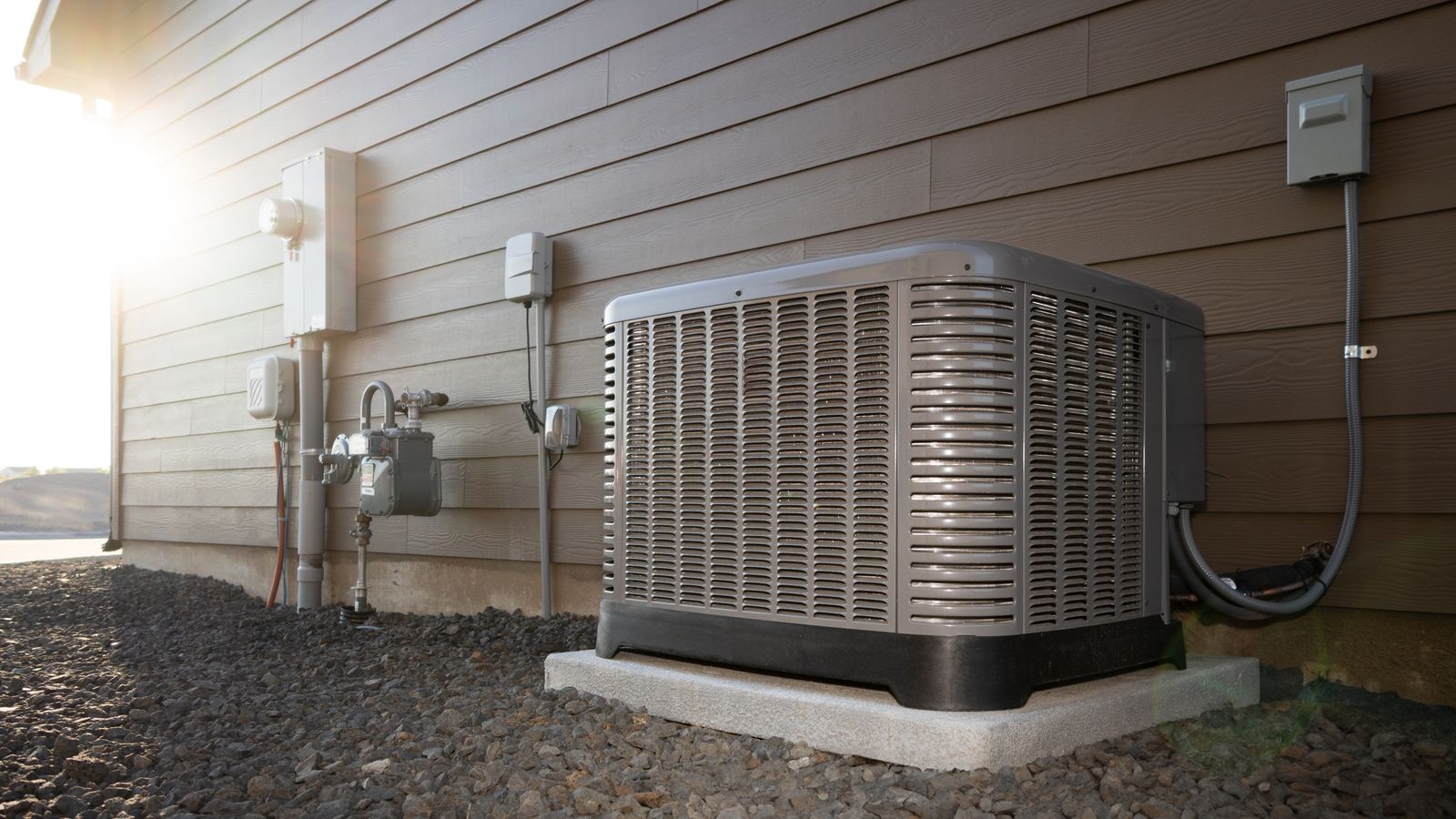 An AC unit in the shade next to a house with the bright sun in the background