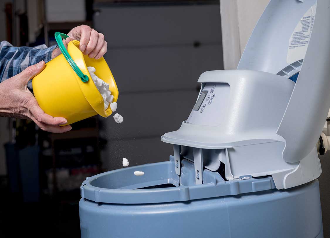 Salt pellets are poured form a yellow bucket into a blue water softener
